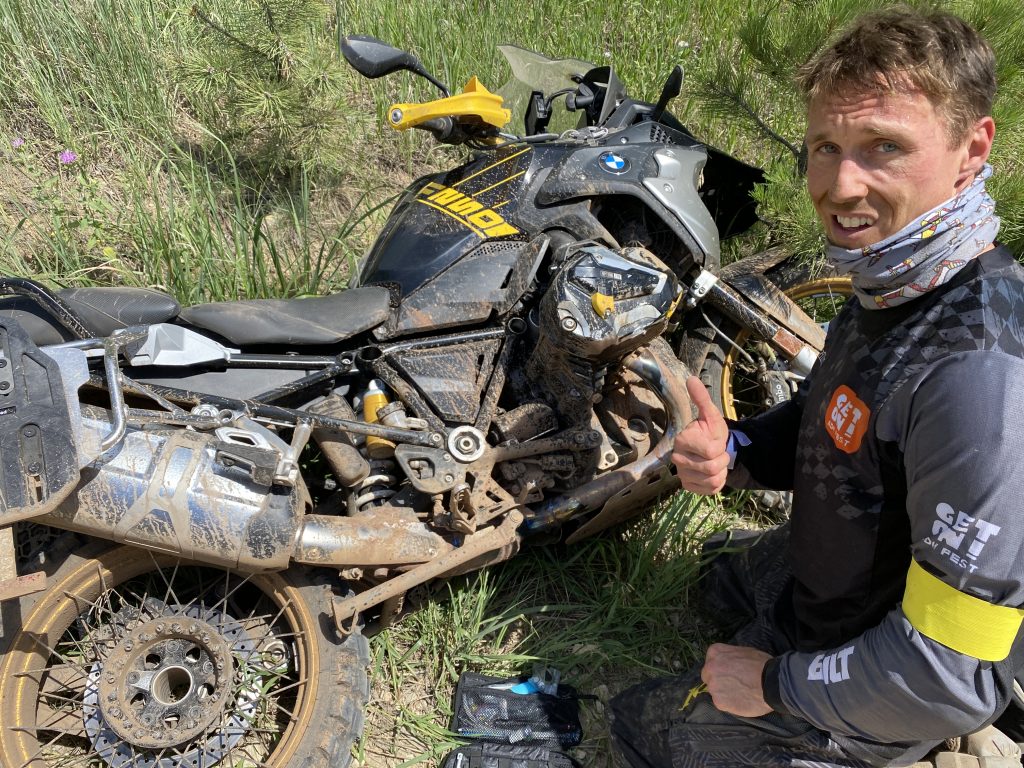 Adventure rider giving thumbs up next to a muddy BMW GS motorcycle after an off-road challenge.